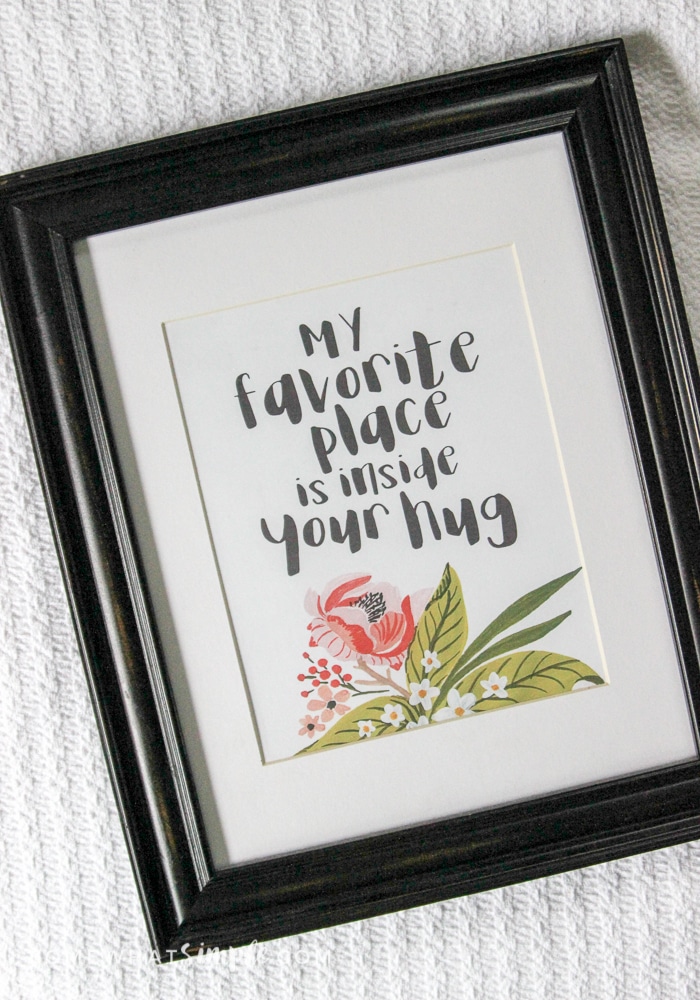 A black framed printable of floral and greenery with the saying, \"My Favorite Place in Inside Your Hug\"