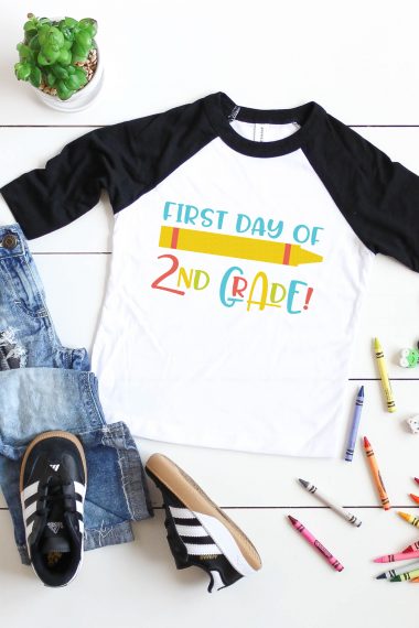 A potted succulent, a pair of black tennis shoes, a pair of blue jeans, scattered crayons and a black and white baseball style shirt with an image of a crayon and the saying "First Day of 2nd Grade"