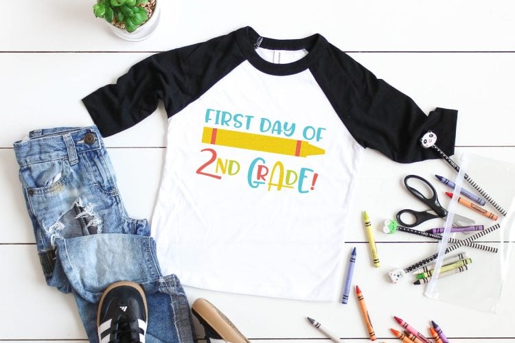 A potted succulent, a pair of black tennis shoes, a pair of blue jeans, scattered crayons and a black and white baseball style shirt with an image of a crayon and the saying \"First Day of 2nd Grade\"