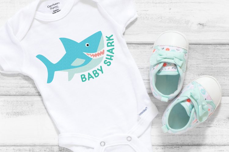 A pair of cute baby tennis shoes and a white onesie with an image of a blue shark on it and the saying, \"Baby Shark\"