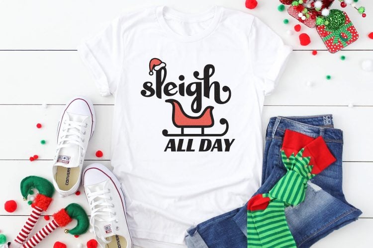 A pair of blue jeans, tennis shoes and green striped elf socks, along with a white t-shirt decorated with the saying, \"Sleigh All Day\" and a sleigh and a Santa hat