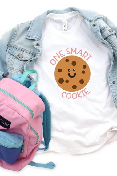 A pink, blue and green colored school backpack next to a blue jean jacket and a white t-shirt with an image of a smiling chocolate chip cookie with the saying, "One Smart Cookie"