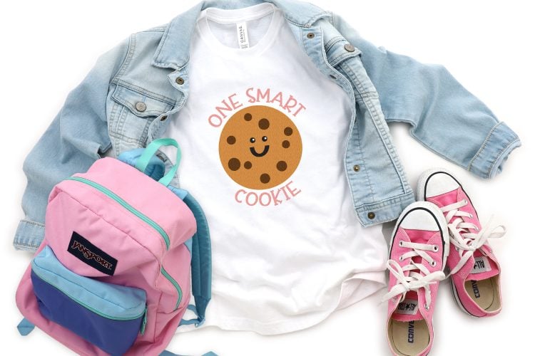 A pair of pink tennis shoes, a pink, blue and green colored school backpack next to a blue jean jacket and a white t-shirt with an image of a smiling chocolate chip cookie with the saying, \"One Smart Cookie\"