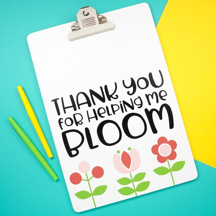 A yellow piece of paper and a green and yellow marker next to a clipboard decorated with flowers and the saying, "Thanks you for Helping Me Bloom"