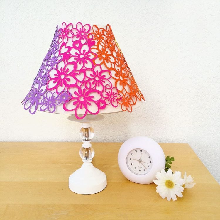 Sitting on a table is a daisy flower, an alarm clock and a lamp with a lacy flower lampshade