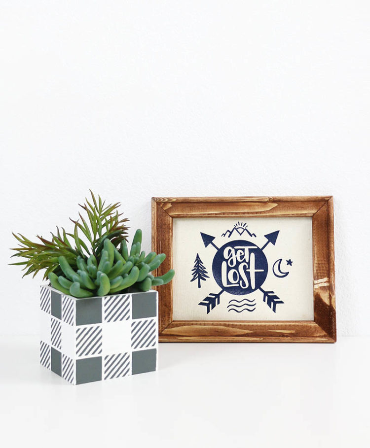 Succulents in a gray and white checkered pot along-side of a wooden framed sign that says, \"Get Lost\"