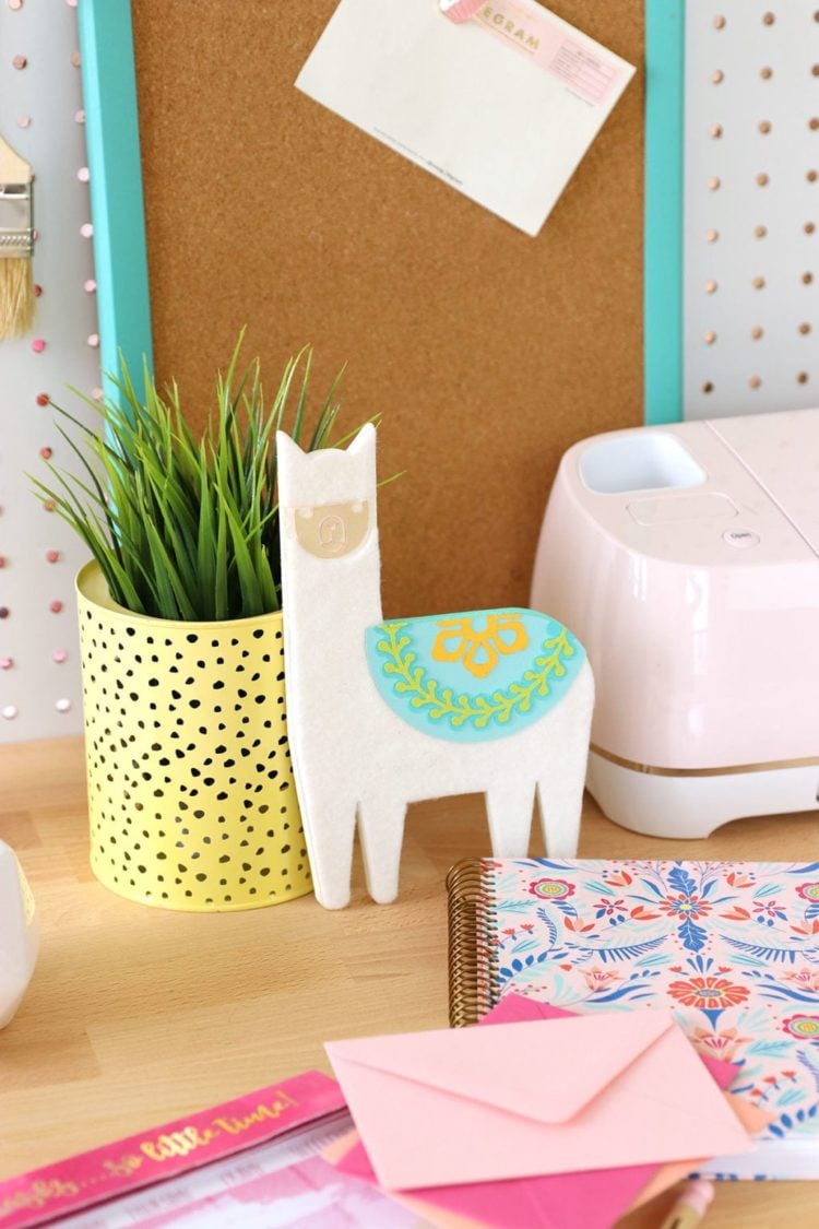 A desk containing a potted plant, a Cricut machine, a journal, an envelope and a figurine of a llama