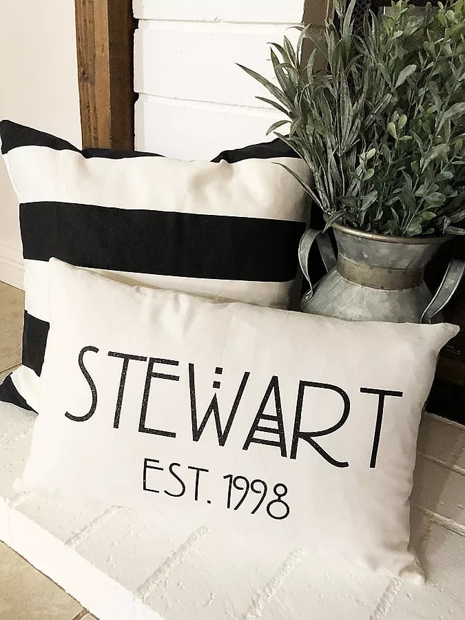 A tin vase filled with greenery, a cream colored and black striped pillow and a cream-colored pillow with a personalized name on it