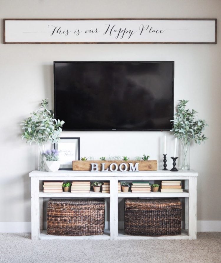 A living room filled with furniture and a flat screen TV and a large wooden sign hanging above the TV that says, \"This is our Happy Place\"