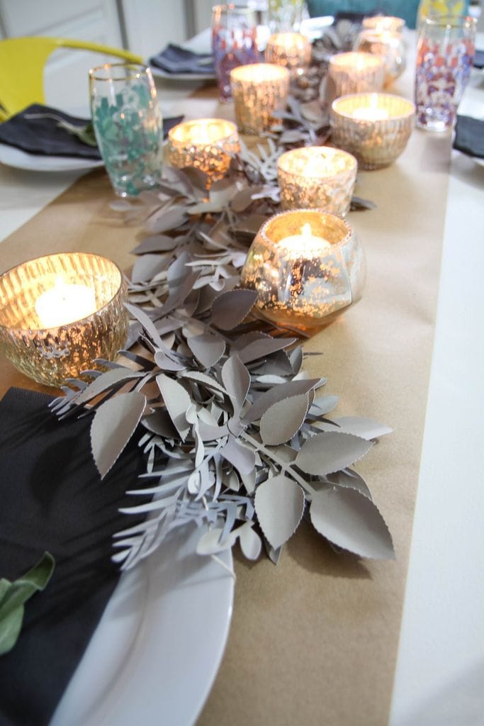 A table runner decorated with paper cutout greenery, candles and place settings