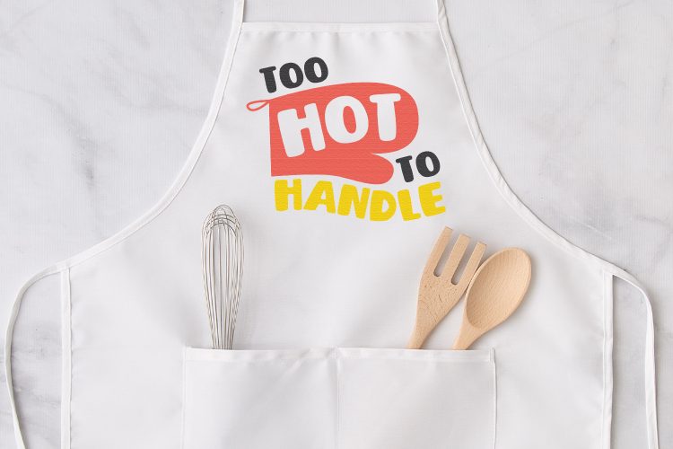 Close up of a kitchen apron with a whisk in one pocket and a wooden fork and spoon in the other pocket.  Apron has a design on it of a red oven mitt and the words \"Too Hot to Handle\" on it