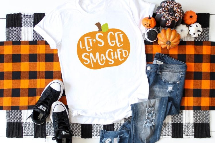 A close-up image of Halloween decor around a pair of blue jeans, a pair of tennis shoes and a white t-shirt that is decorated with an orange pumpkin that says, \"Let\'s Get Smashed\"