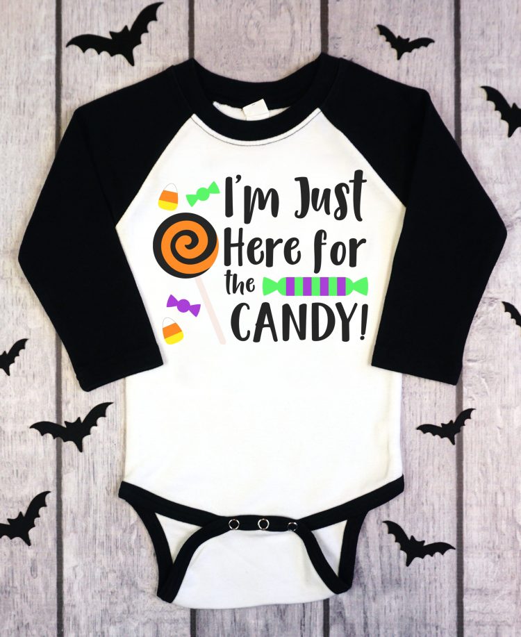 A onesie with black sleeves and white body that is decorated with the Halloween saying, \"I\'m Just Here for the Candy\". Shirt is surrounded by black paper cut out bats. 