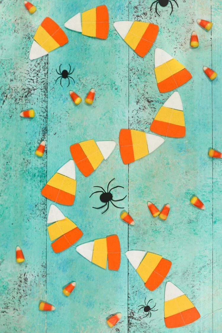 A candy corn garland cut out of felt surrounded by black cut out paper spiders and real candy corn candy