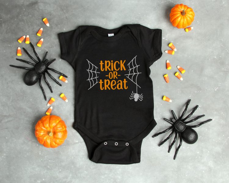 Halloween decor of black spiders, orange mini pumpkins, candy corn candy surrounding a black onesie designed with a spider and a web on it and with the saying, \"Trick or Treat\"