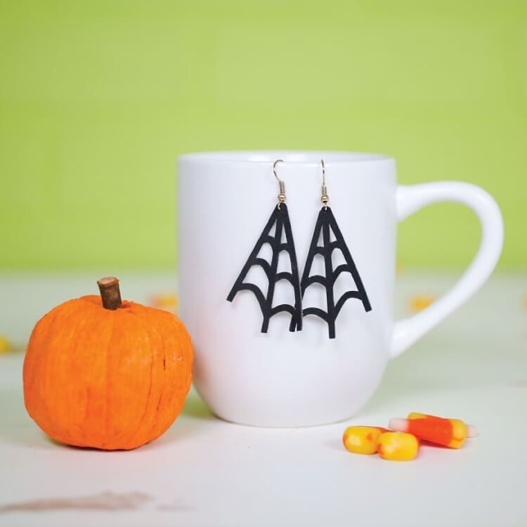 A small orange pumpkin, some candy corn candy and a white coffee mug with black spider web earrings hanging on it