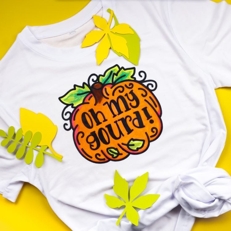 White t-shirt with a design of a pumpkin that says, "Oh My Gourd".  Several paper-cut leaves lay on top of the shirt.