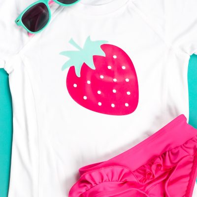 Pink swimming bottoms and a pair of sunglasses lying on top of a white t-shirt decorated with a pink strawberry