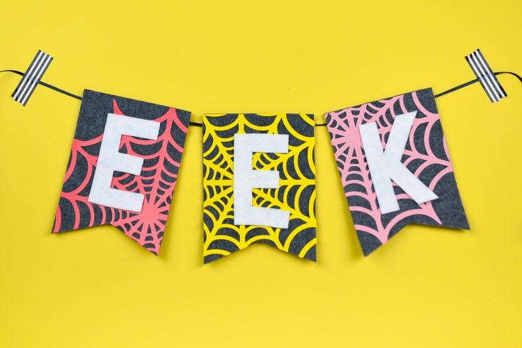 A close up of three pieces of a Halloween banner made of grey felt hanging on a bright yellow wall.  One piece of the banner has a felt orange spider web, one with a yellow spider web and that third one with a pink spider web and the banner spells out the word, "EEK" with all letters being cut out of white vinyl.