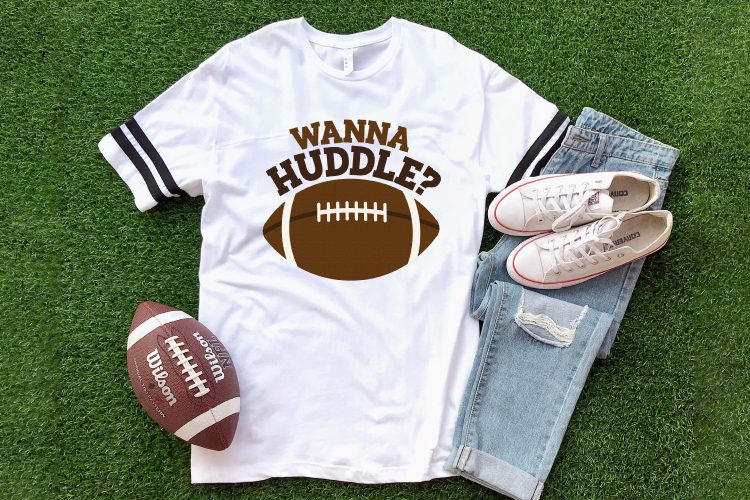 A football, a pair of blue jeans, a pair of tennis shoes all lying on green turf along with a white t-shirt designed with a football and the saying, \"Wanna Huddle?\"