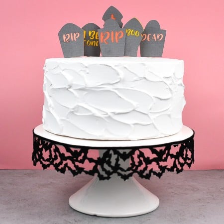 A white frosted cake sitting on a black and white cake pedestal and the cake is decorated with a graveyard topper