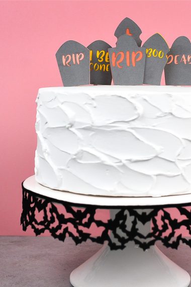 A white frosted cake sitting on a black and white cake pedestal and the cake is decorated with a graveyard topper