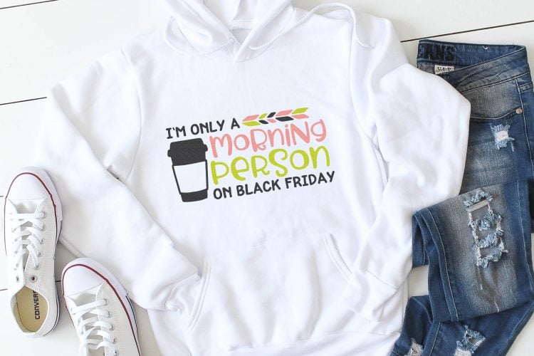 A pair of blue jeans, tennis shoes and a white hooded sweatshirt that is decorated with a coffee tumbler and a saying that says, \"I\'m Only a Morning Person Black on Friday\"