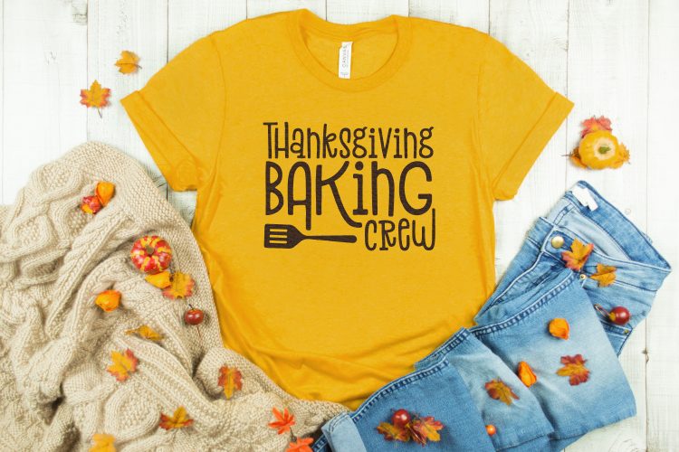 A heavy sweater, a pair of blue jeans and a pink t-shirt surrounded by fall decor and the shirt has the design of a spatula and text that says, \"Thanksgiving Baking Crew\"