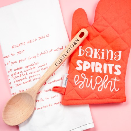 Close up of a pink oven mitt that says, "Baking Sprits Bright" and a wooden spoon that says, "Ellen's Kitchen" and a white kitchen towel decorated with a recipe