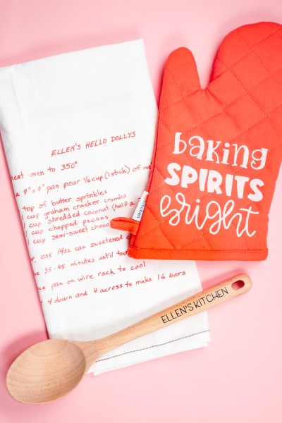 A pink oven mitt that says, "Baking Sprits Bright" and a wooden spoon that says, "Ellen's Kitchen" and a white kitchen towel decorated with a recipe