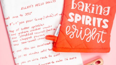 A pink oven mitt that says, "Baking Sprits Bright" and a wooden spoon that says, "Ellen's Kitchen" and a white kitchen towel decorated with a recipe