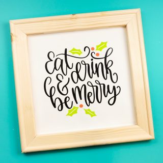 A close up of a wooden framed sign against a dark aqua painted wall that says, "Eat Drink and be Merry"