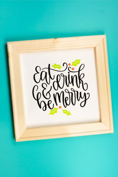 A wooden framed sign against a dark aqua painted wall that says, "Eat Drink and be Merry"