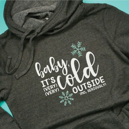 A dark grey hooded sweatshirt decorated with greenish colored snowflakes and the saying "Baby It's (VERY VERY) Cold Outside (No, Seriously)"