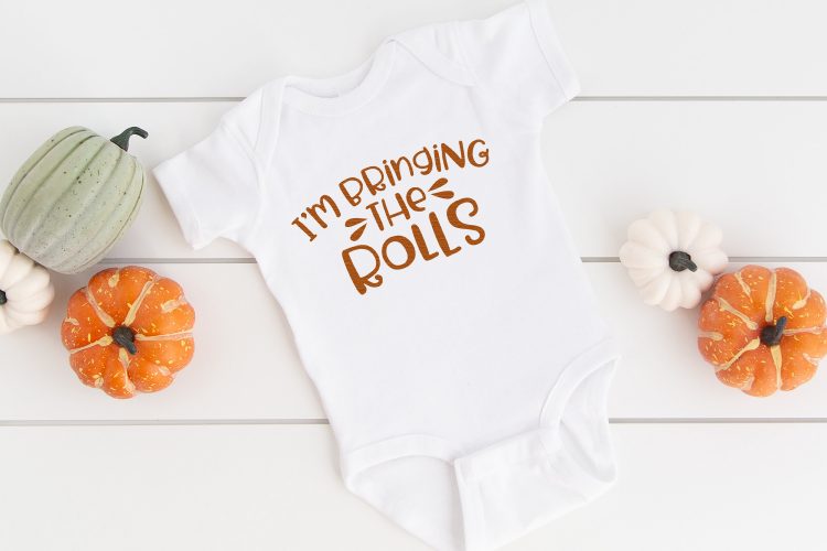 A white onesie surrounded by different colored gourds and the onesie has the saying, \"I\'m Bringing the Rolls\" on it