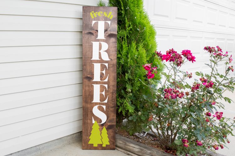 A vertical Christmas sign on the side of a building next to a small tree and a flowering bush. The sign says, \"Fresh Trees\".