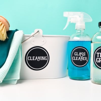 A glass bottle with the label "Tile & Grout" on it, a spray bottle with the label "Glass Cleaner", and a bucket labeled "Cleaning" with a scrub brush in it and cleaning rags hanging over it