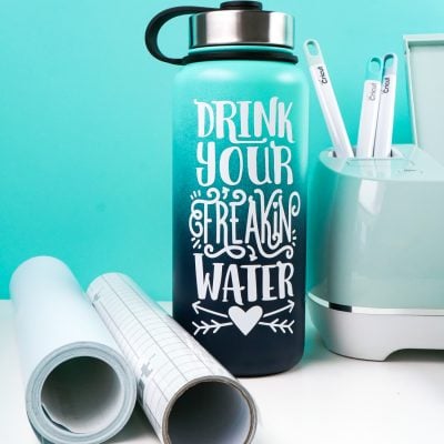 A Cricut machine along with vinyl and transfer tape and a water bottle with the saying "Drink Your Freaking Water" on it.