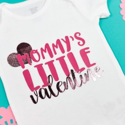 A white onesie with a heart and quote "Mommy's Litte Valentine".  Onesie is surrounded by pink doilies and cutout hearts.