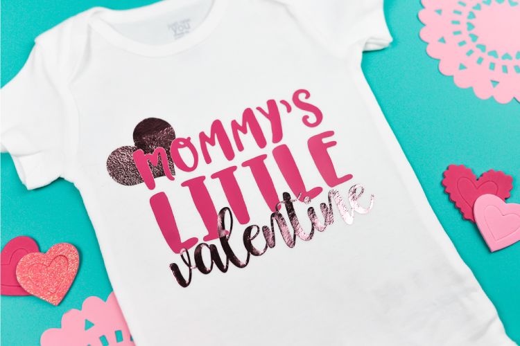 A white onesie with a heart and quote \"Mommy\'s Litte Valentine\".  Onesie is surrounded by pink doilies and cutout hearts.
