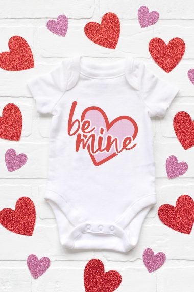 A white onesie surrounded by cut out hearts and the saying on the onesie "Be Mine" on top of a pink heart outlined in red