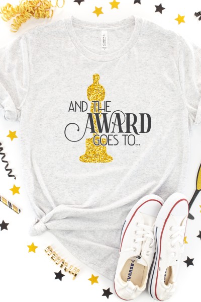 A cream-colored t-shirt surrounded by a pair of tennis shoes and confetti.  The shirt has an image of the Oscars Award in gold glitter vinyl and text in black vinyl that says, "And The Award Goes To"