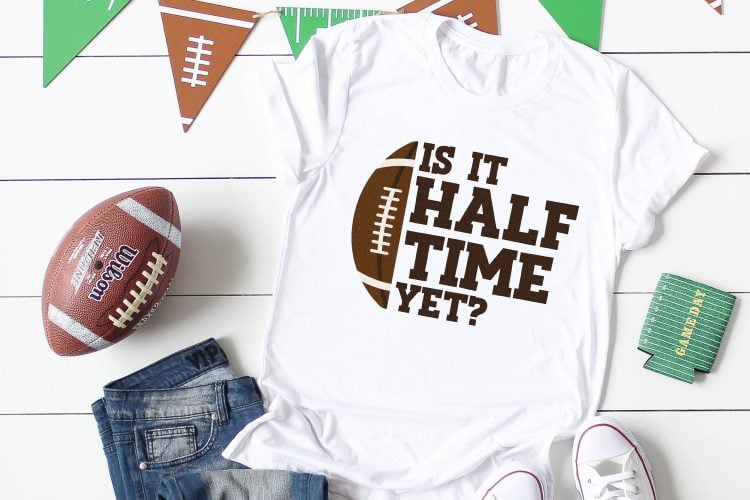 A pair of blue jeans, tennis shoes, football, football themed banner, a football themed koozie and a white shirt with picture of a half of a football and the saying, \"Is It Half Time Yet?\"