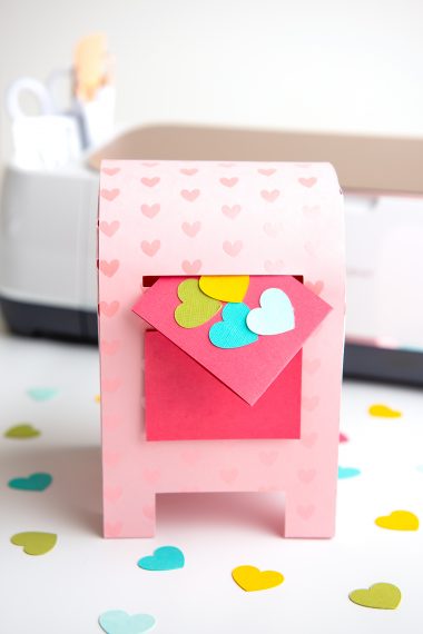 A paper Valentine's Day Mailbox with Cricut machine in background and colored hearts scattered around the mailbox