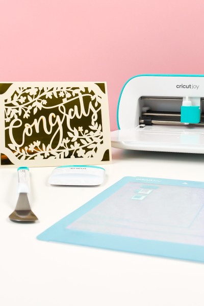 Cricut Joy machine is pictured with a 'Congrats' greeting card and Cricut mat and tools