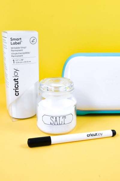 A Cricut Joy pictured with a Cricut Joy black pen, a box of Cricut Joy Smart Label vinyl and a jar that contains a label that says 'Salt'.