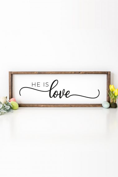 A framed sign with a white background with quote in black vinyl that says, "He is Love".  Frame is set among Easter decor.