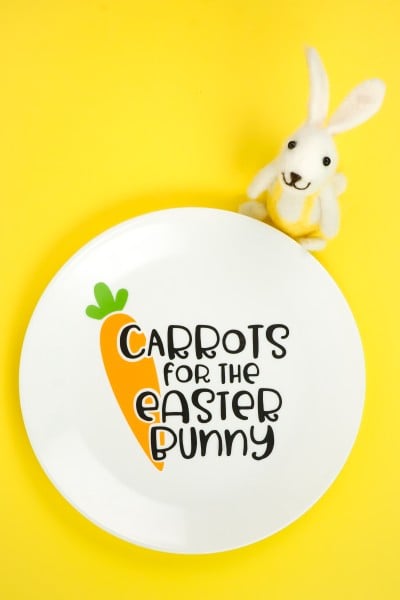 A bunny figurine standing next to a white dinner plate that has a design of a carrot and the saying, "Carrots for the Easter Bunny" on it