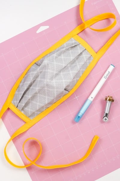 Picture of a completed face mask lying on a pink FabricGrip Cricut mat along with the Cricut rotary blade tool and knife
