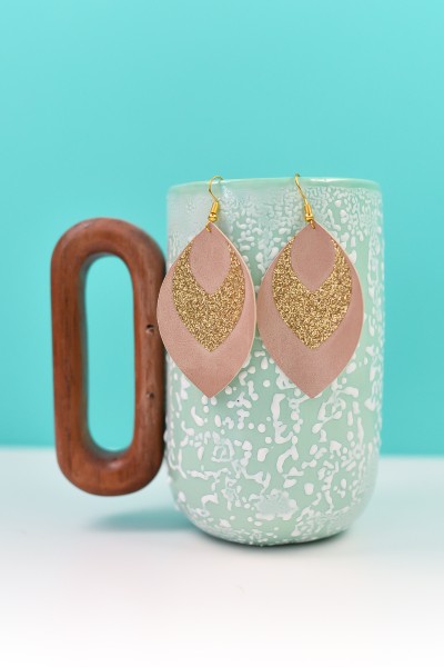Glitter and suede Cricut earrings hanging over an aqua blue coffee mug that has a wooden handle
